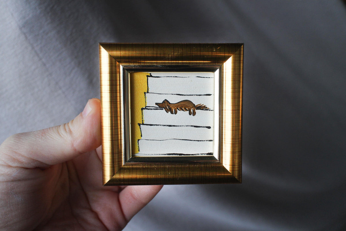 Small framed artwork with a gold frame held by a hand against a gray background