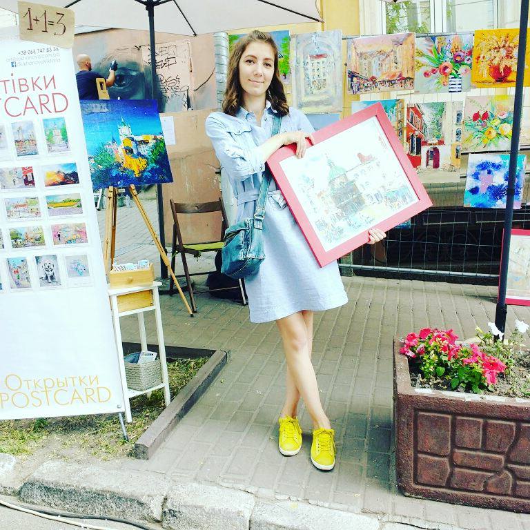 A woman stands outdoors holding a framed artwork with a pink border. She is wearing a light blue denim jacket and dress with bright yellow shoes, surrounded by art displays, posters, and flowers on a city sidewalk.