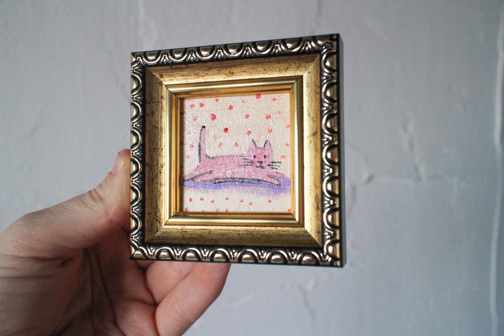 Small painting of a pink cat on a polka dot background in a decorative gold frame held by a hand against a plain background.