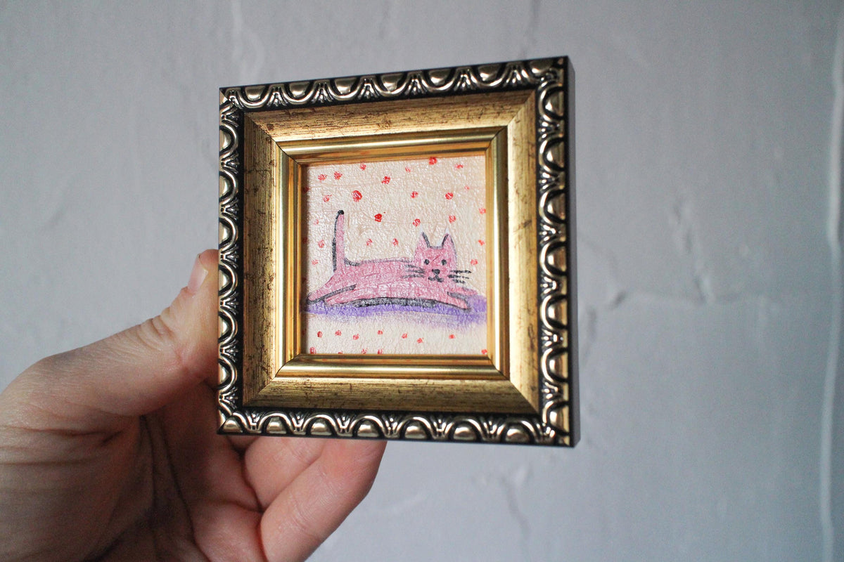 Small painting of a pink cat on a polka dot background in a decorative gold frame held by a hand against a plain background.