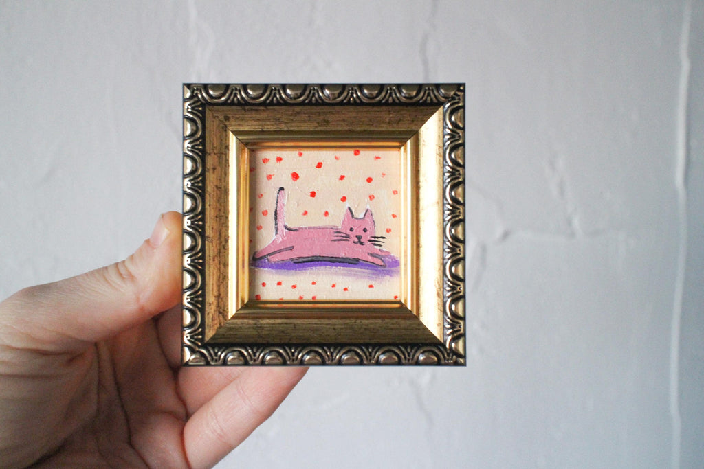 Small painting of a pink cat on a polka dot background in a decorative gold frame held by a hand against a plain background.