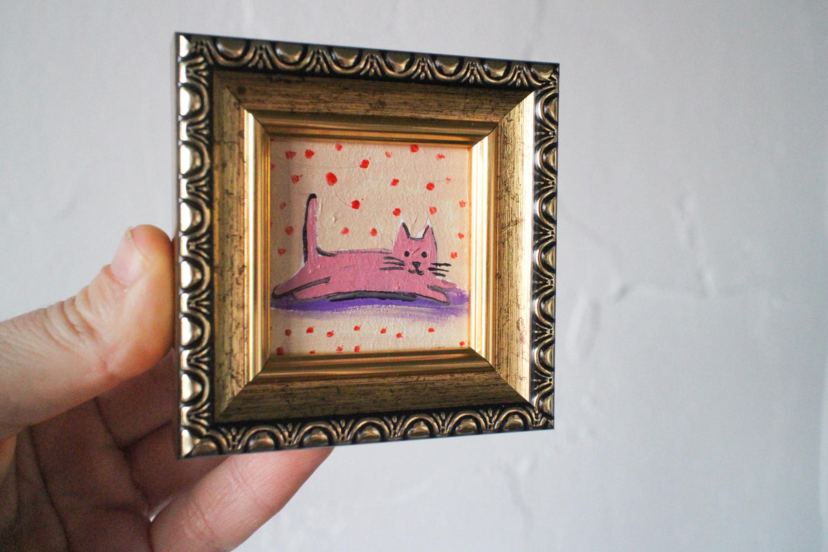 Small painting of a pink cat on a polka dot background in a decorative gold frame held by a hand against a plain background.