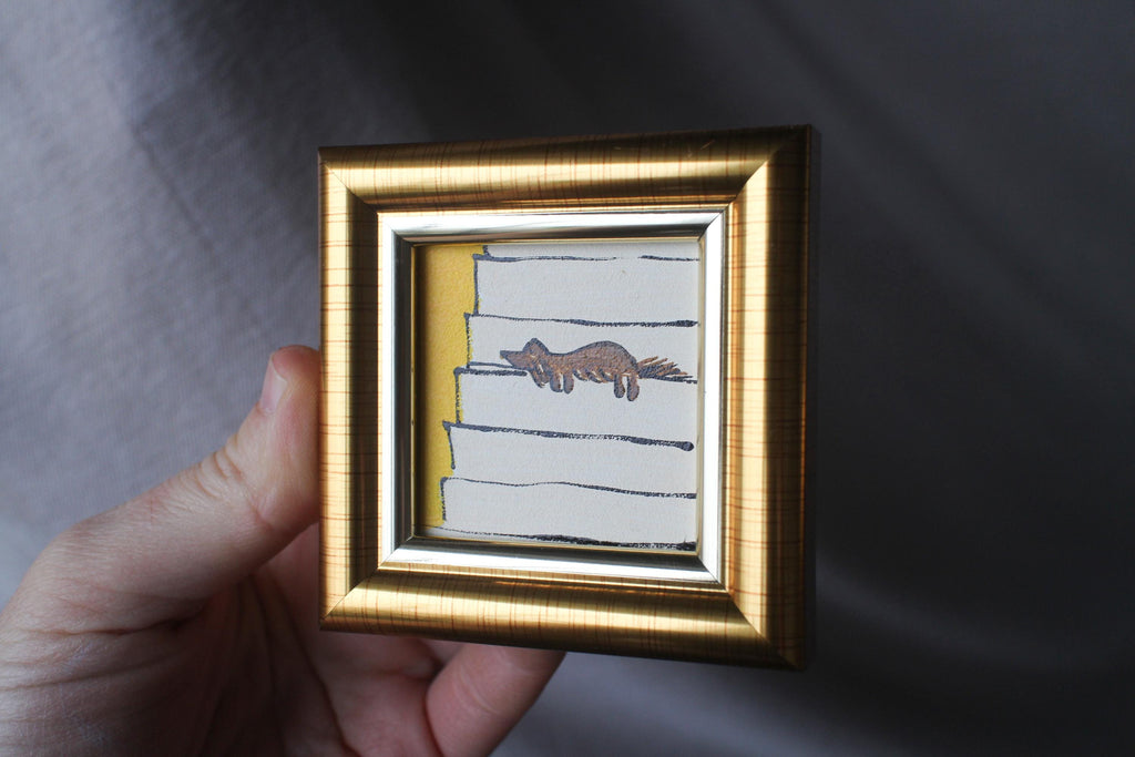 Small framed artwork with a gold frame held by a hand against a gray background