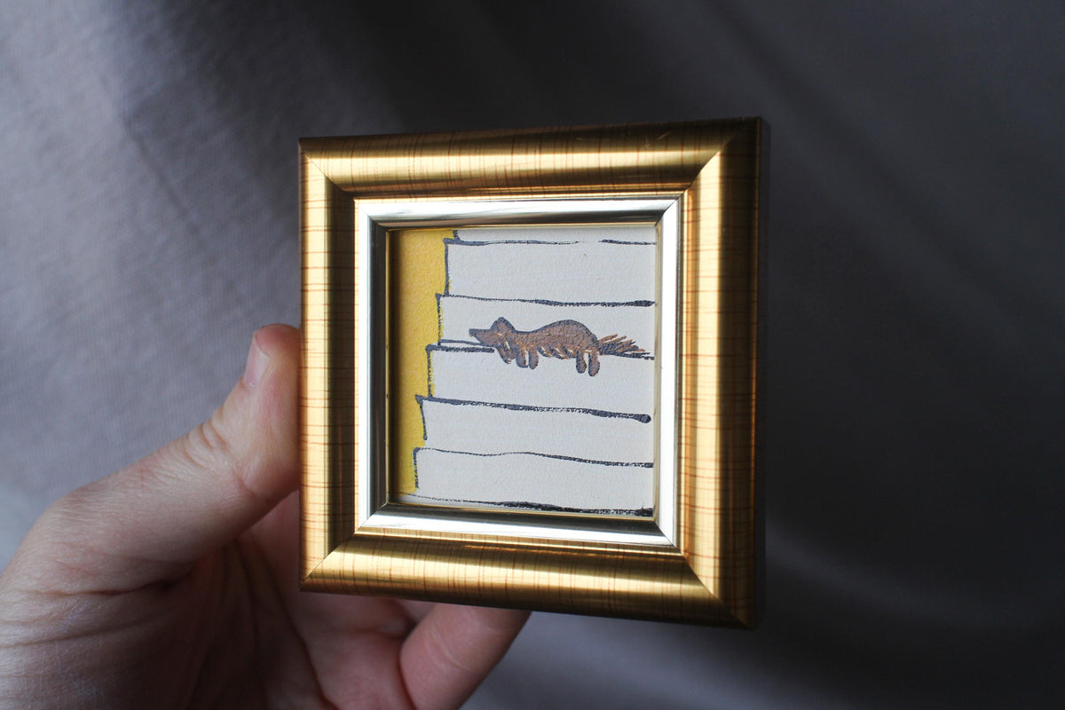 Small framed artwork with a gold frame held by a hand against a gray background