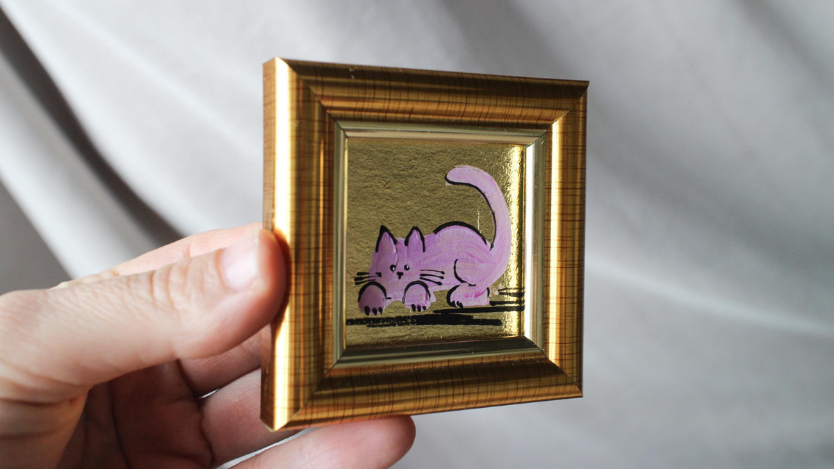 Small gold-framed picture with a purple cat illustration held by a hand against a neutral background