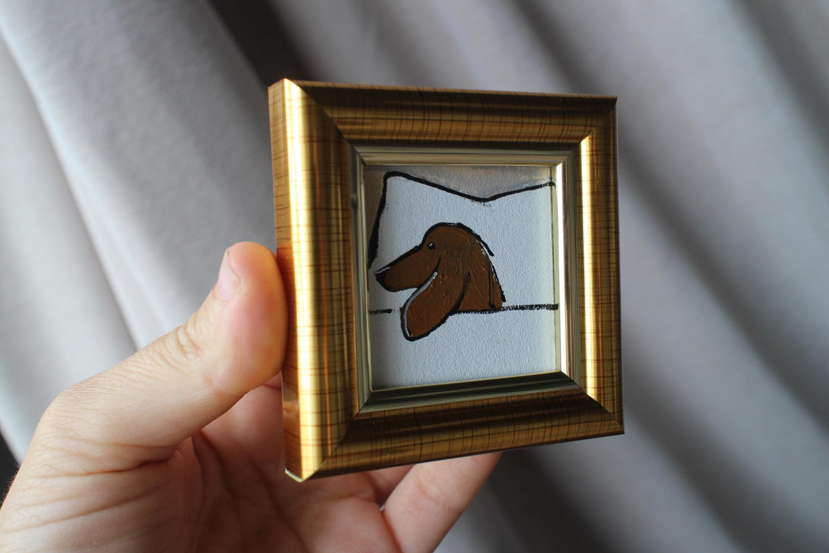 Small framed artwork of a dog's face held by a hand against a neutral background