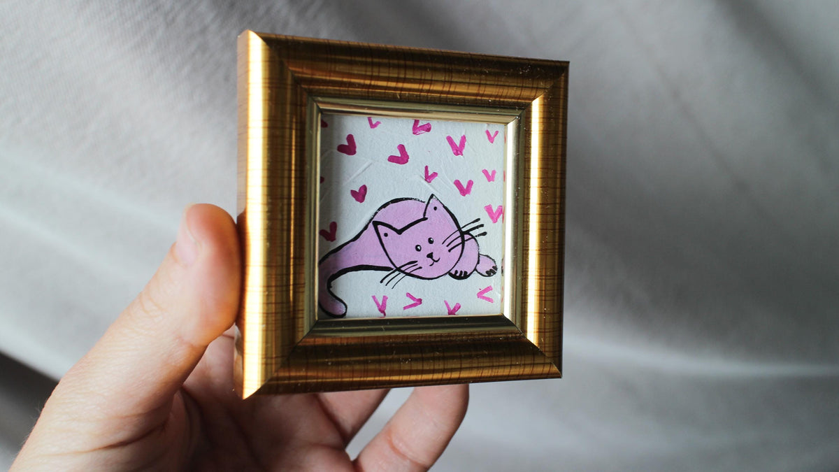 Hand holding a small gold-framed picture with a pink cat illustration on a white background.