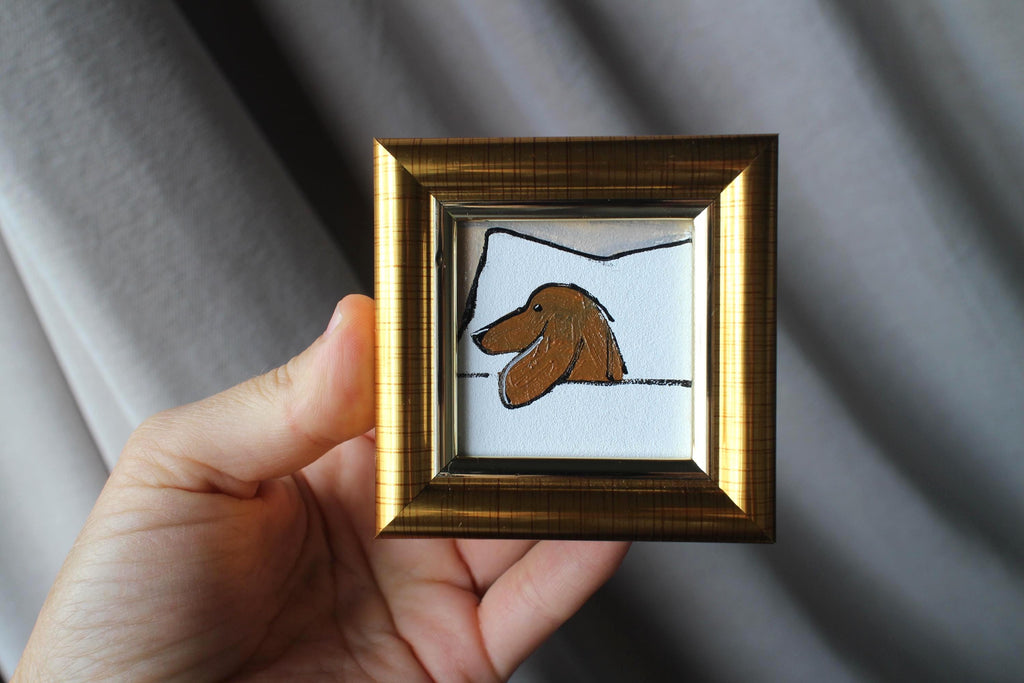 Hand holding a small framed drawing of a dog's face against a gray background