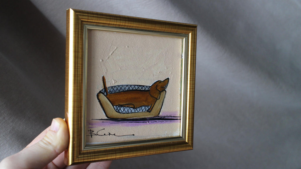 Framed artwork of a dog in a basket held by a hand against a gray background
