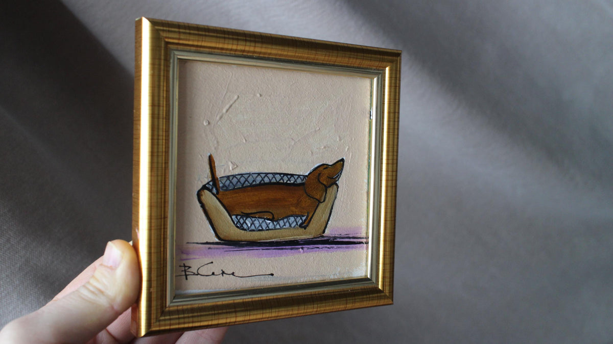 Framed artwork of a dog in a basket held by a hand against a gray background