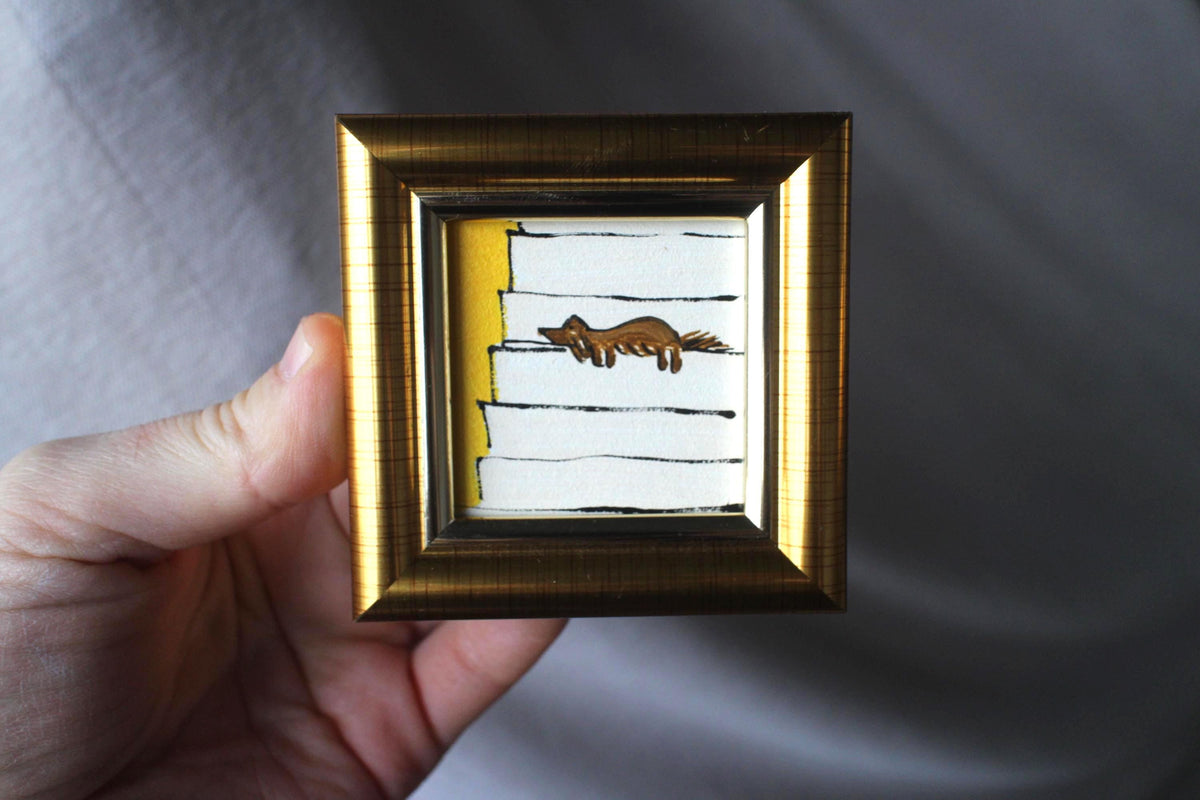 Small framed artwork with a gold frame held by a hand against a gray background