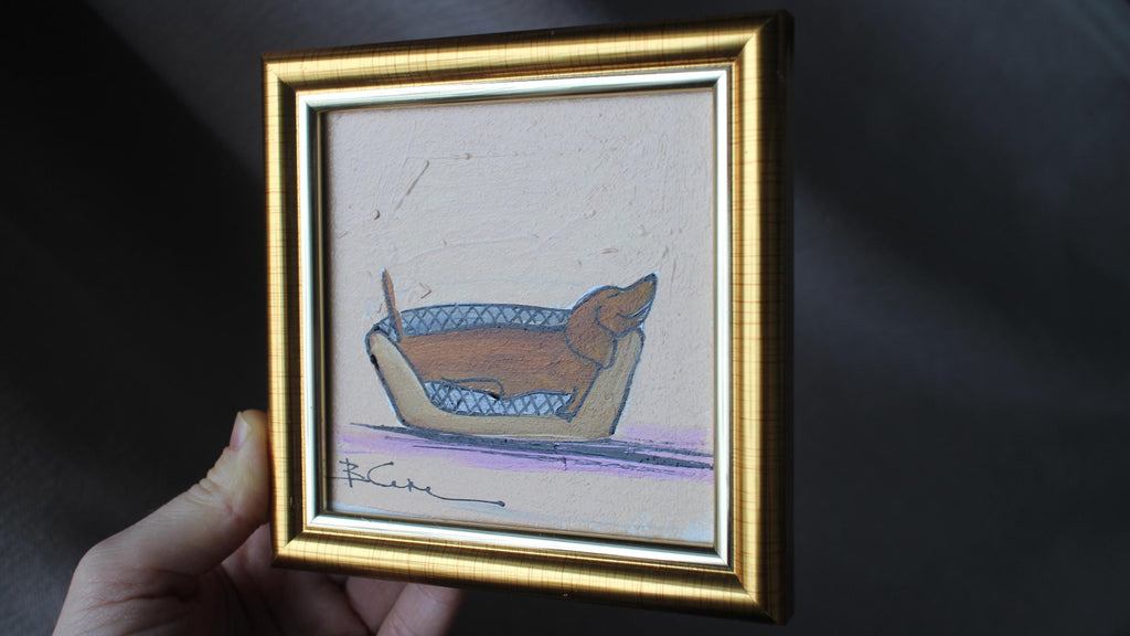 Framed drawing of a dog in a blue bed held by a hand against a dark background