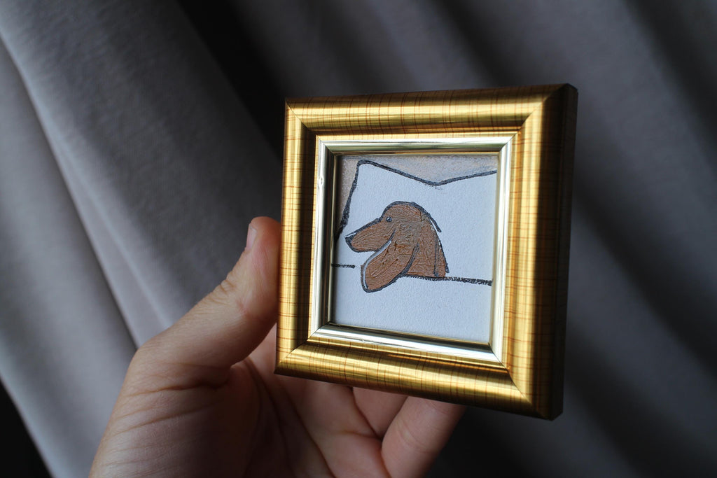Hand holding a small gold-framed artwork of a dog's face against a dark background
