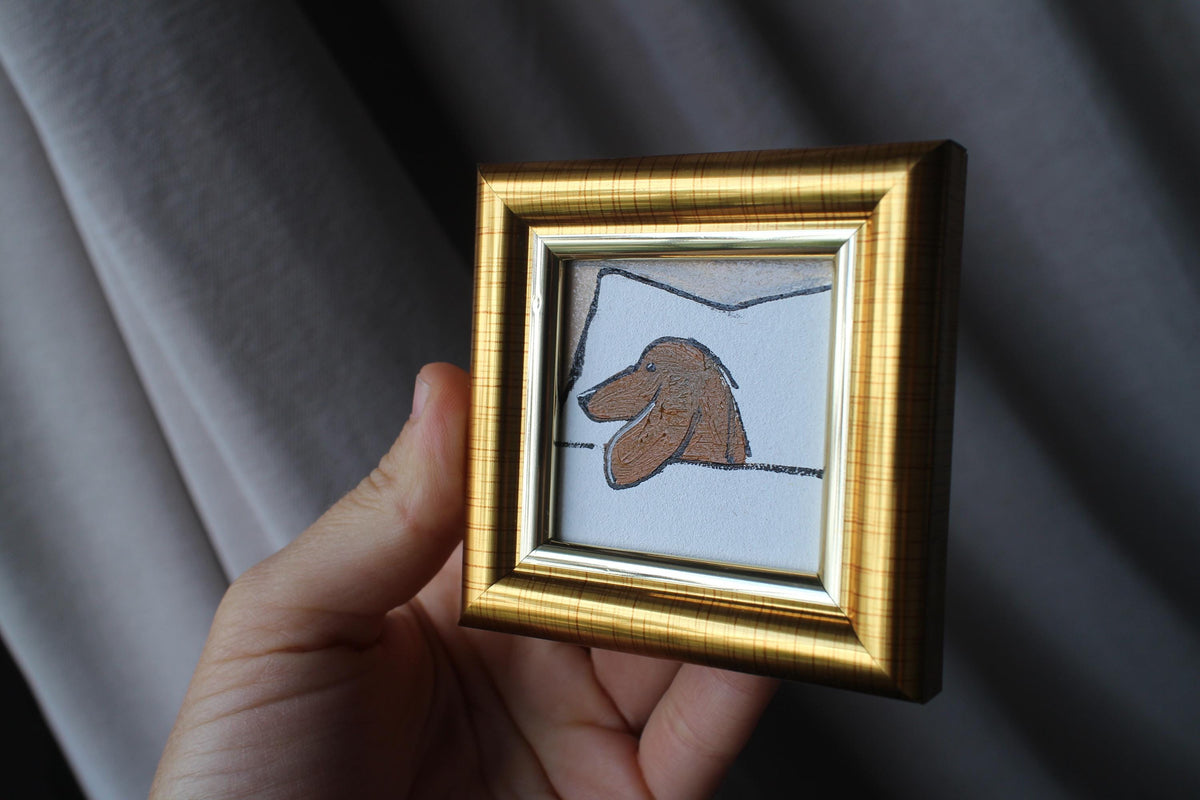 Hand holding a small gold-framed artwork of a dog's face against a dark background