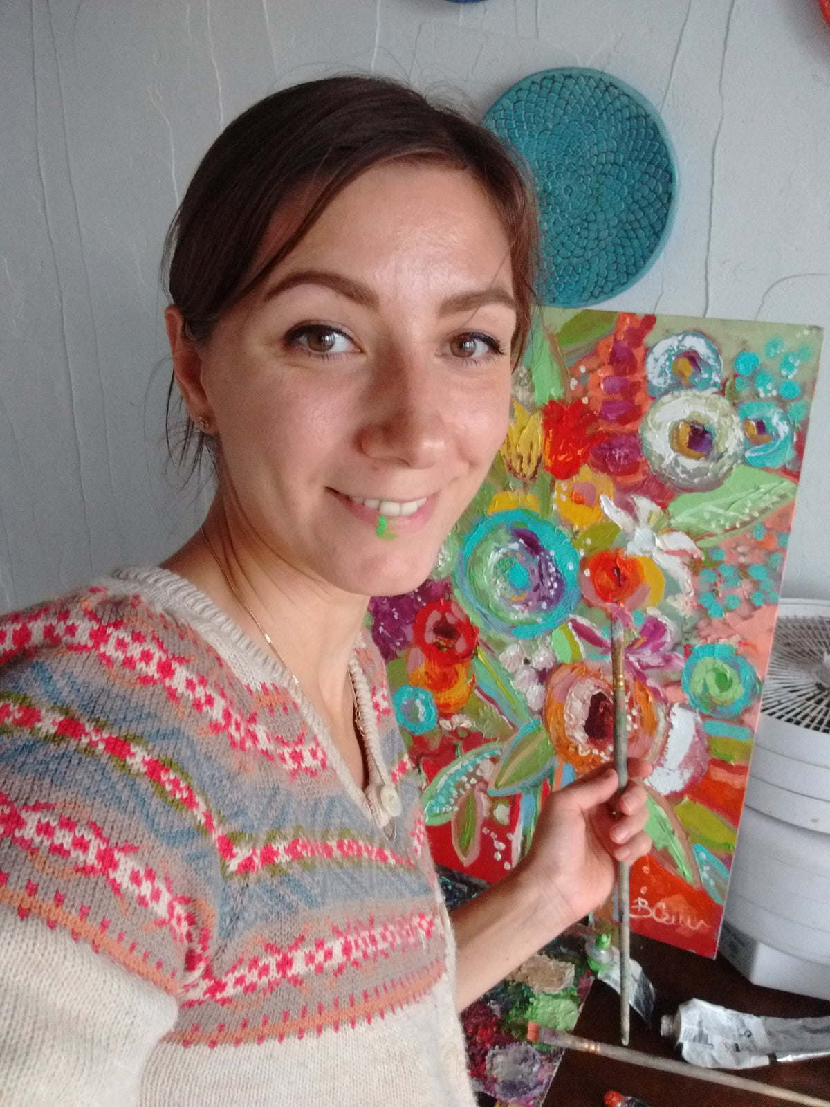 A woman holding a paintbrush in front of a colorful abstract painting, wearing a cream knit sweater with pink, red, and blue geometric patterns.