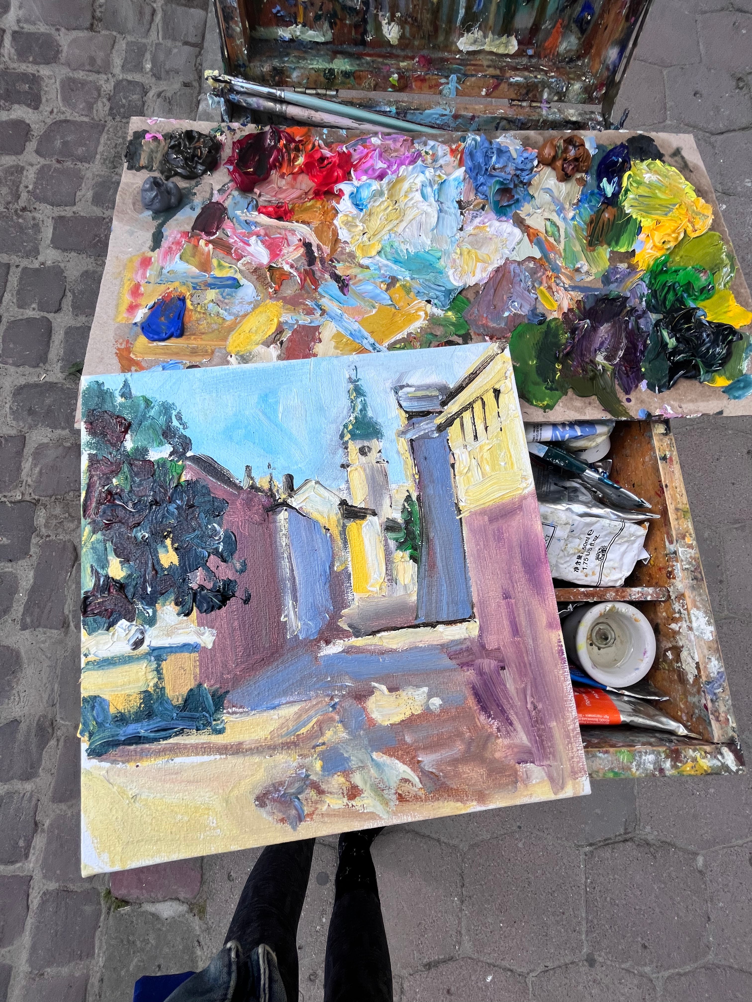 An open artist palette filled with mixed paint colors and brushes, with a painted canvas showing an impressionistic street scene placed in front, photographed outdoors on a cobblestone surface.