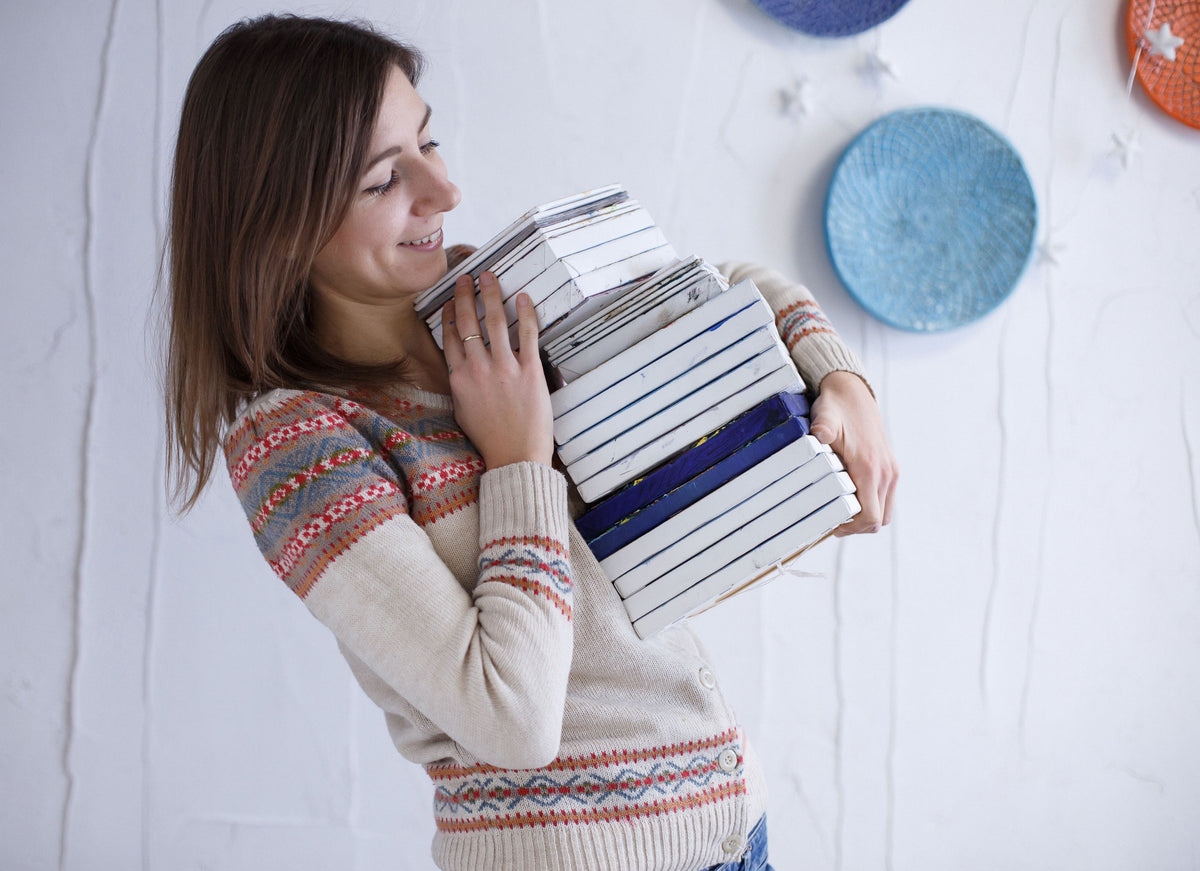 A woman with long brown hair smiles while carrying a large stack of white books or boxes with blue stripes, wearing a cream knit sweater with colorful Fair Isle patterns on the shoulders and hem, set against a white wall with decorative blue plates.