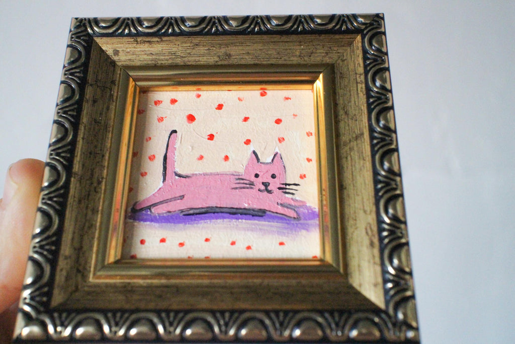 Small painting of a pink cat on a polka dot background in a decorative gold frame held by a hand against a plain background.