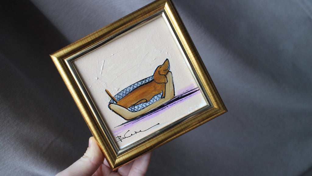 Small framed artwork of a dog in a boat held by a hand on a dark fabric background