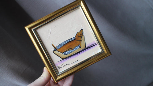 Small framed artwork of a dog in a boat held by a hand on a dark fabric background