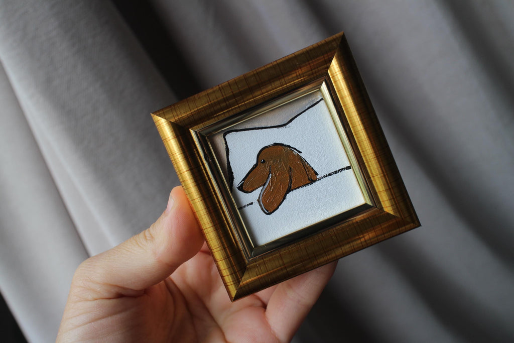 Small framed artwork of a dog held by a hand against a gray background