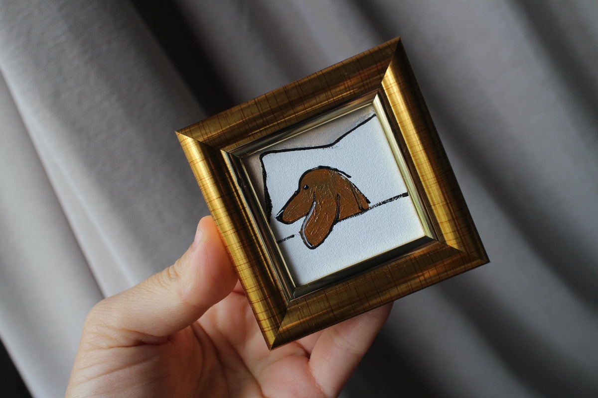 Small framed artwork of a dog held by a hand against a gray background