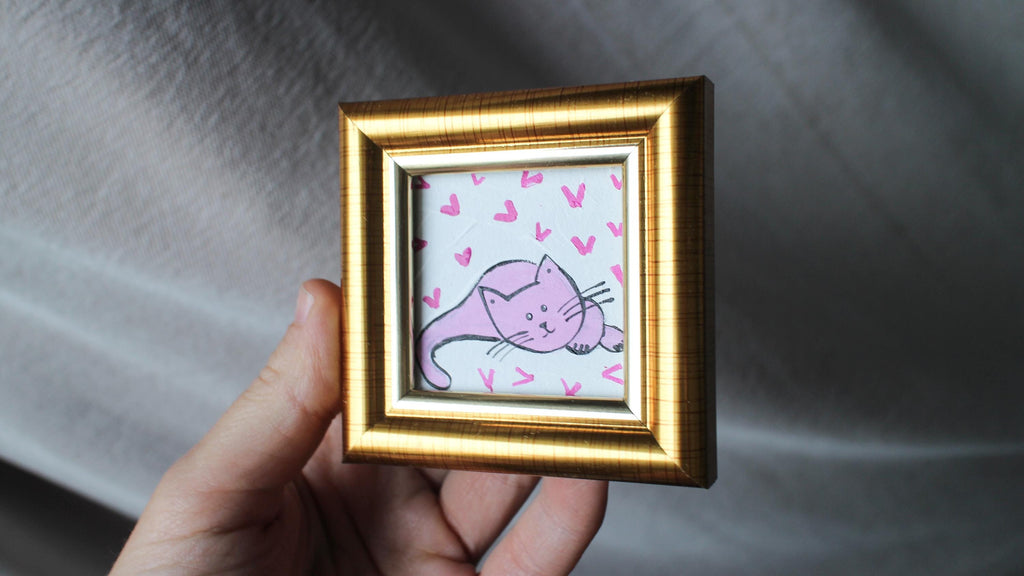 Hand holding a small gold-framed picture with a pink cat illustration on a white background.