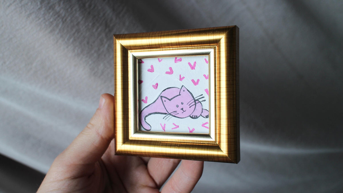 Hand holding a small gold-framed picture with a pink cat illustration on a white background.