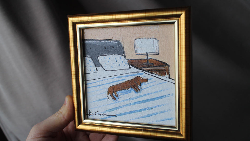 Framed artwork of a dog on a bed held by a hand against a dark background
