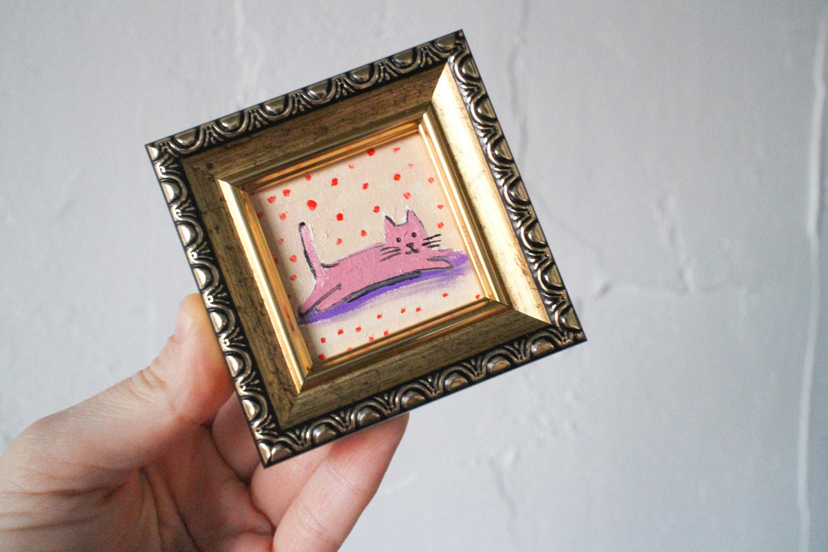 Small painting of a pink cat on a polka dot background in a decorative gold frame held by a hand against a plain background.