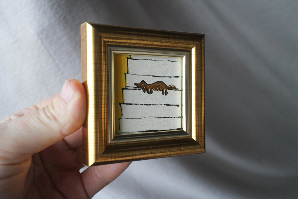 Small framed artwork with a gold frame held by a hand against a gray background