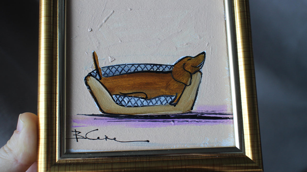 Framed artwork of a dachshund in a basket on a textured beige background.