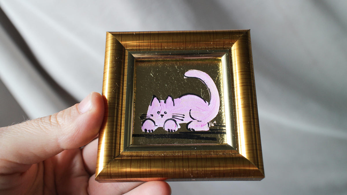 Small gold-framed picture with a purple cat illustration held by a hand against a neutral background