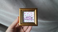 Small framed artwork of a pink cat with hearts held by a hand against a gray fabric background
