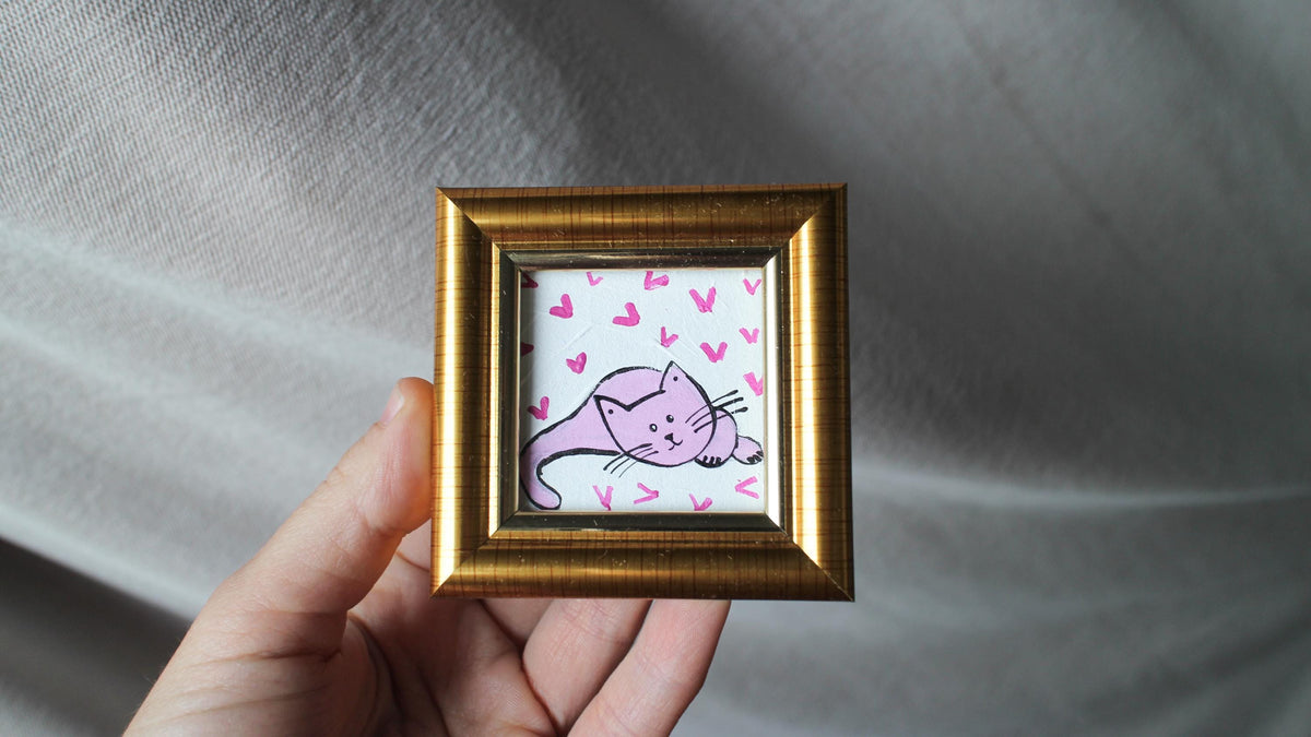 Small framed artwork of a pink cat with hearts held by a hand against a gray fabric background
