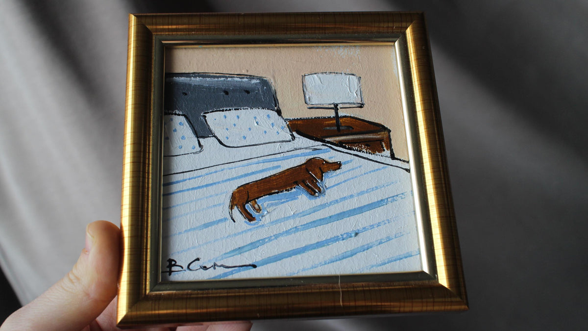 Framed miniature painting of a dog on a bed held by a hand.