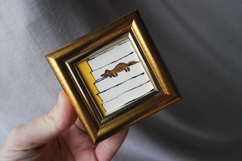 Small framed artwork with a gold frame held by a hand against a gray background